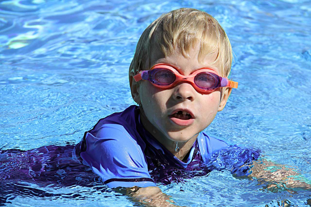 Child swimming in a saltwater swimming pool with red goggles and a blue swim shirt, enjoying a sunny day.